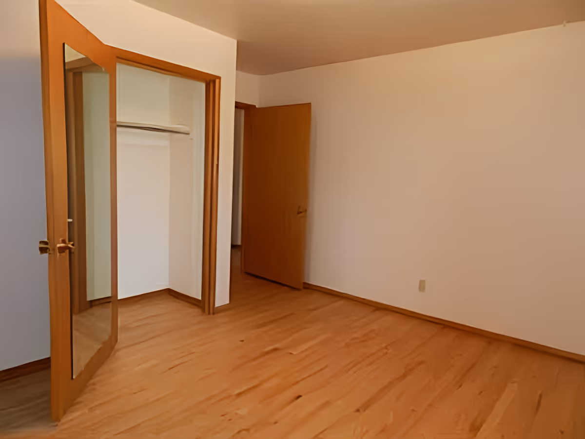Empty bedroom with hardwood floors, an open mirrored closet door and a wooden entry door.