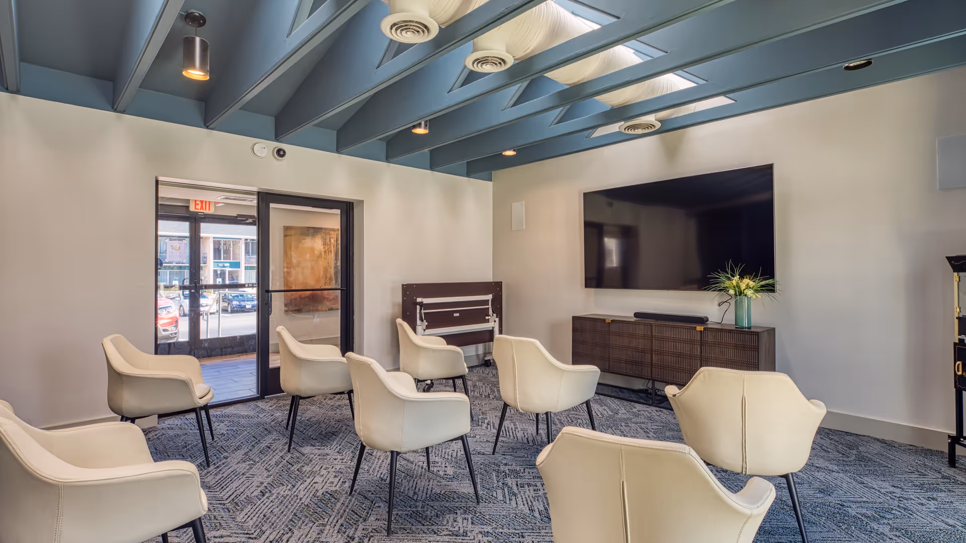 A modern meeting or media room with several cream-colored chairs arranged facing a large flat-screen TV mounted on the wall. The room has a blue ceiling with exposed beams and skylights, a patterned carpet, and a glass door leading outside.