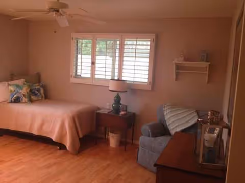A cozy bedroom featuring a bed, nightstand, window with blinds, and blue chair.