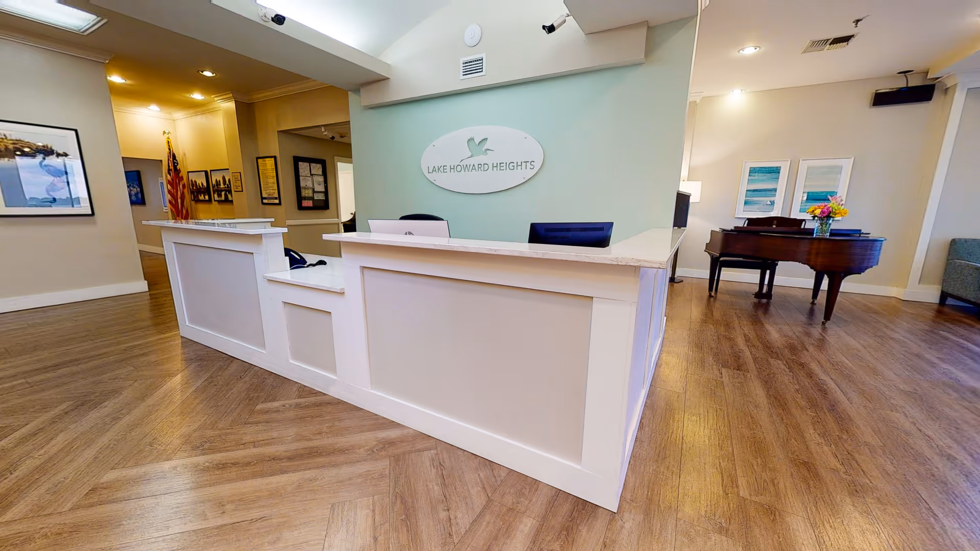 Reception area of Lake Howard Heights facility with a white front desk, a sign on the wall behind the desk displaying the facility name and logo, wooden flooring, an American flag in the hallway, framed pictures on the walls, and a grand piano with a vase of flowers in the background.