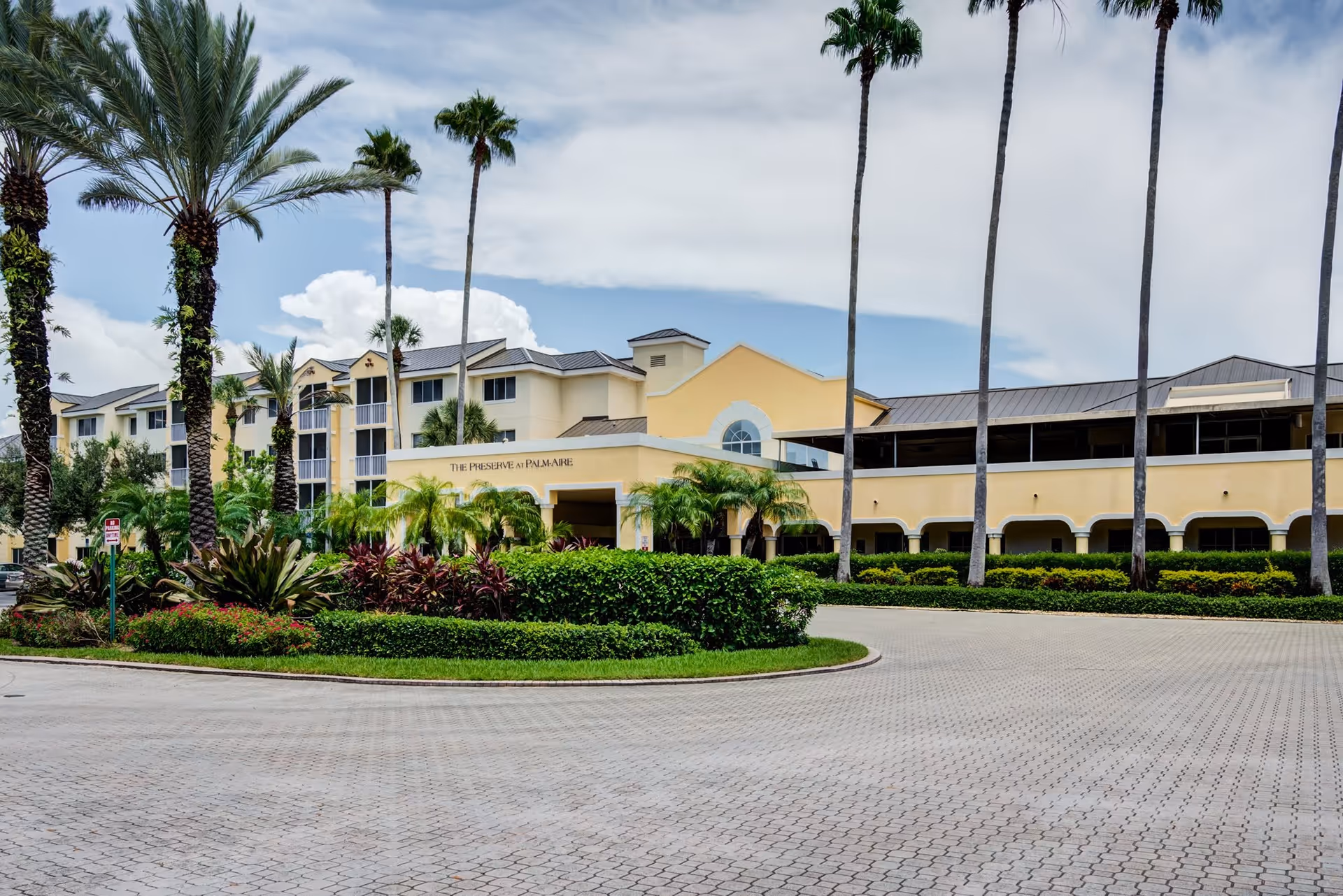 Exterior view of The Preserve at Palm-Aire building with a paved driveway, landscaped greenery, and tall palm trees under a partly cloudy sky.