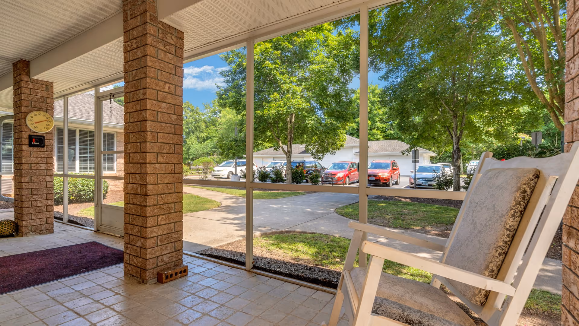 View from a covered porch with brick pillars and a cushioned rocking chair, overlooking a parking area with several cars and green trees under a blue sky.