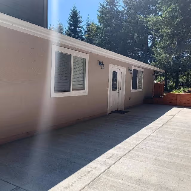 Exterior view of a single-story building with beige walls, two windows with white frames, a white double door, and outdoor wall lights. The building is surrounded by tall evergreen trees and has a concrete patio area in front.