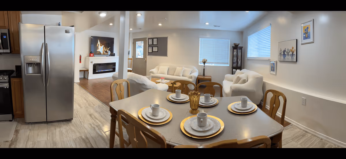 A bright and cozy interior space featuring a dining area with a table set for four with white plates, cups, and gold chargers. Adjacent to the dining area is a living room with white sofas, a coffee table, a fireplace with a TV above it, and windows with blinds letting in natural light. The kitchen area includes a stainless steel refrigerator and wooden cabinets.