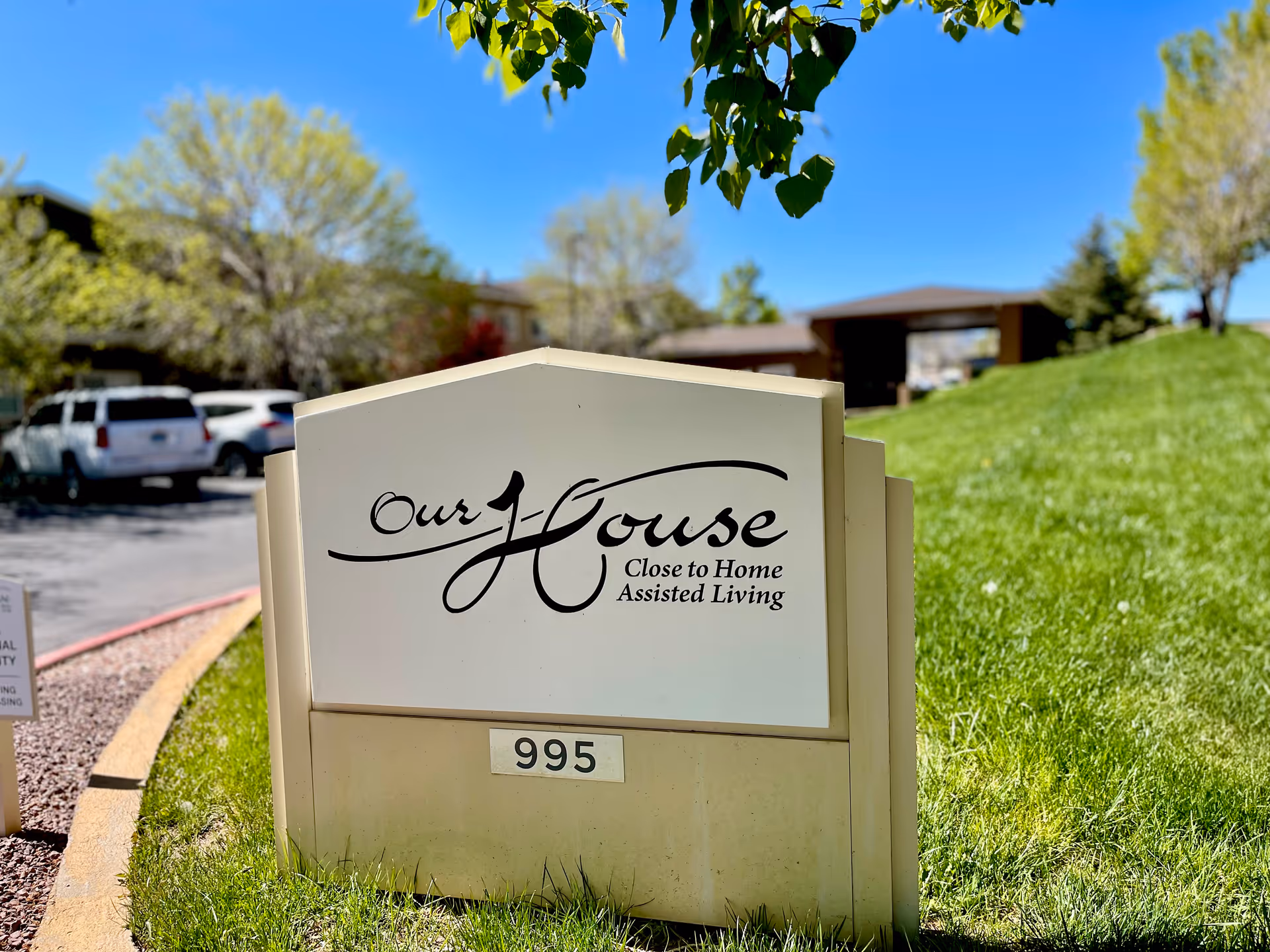 Outdoor view of a sign for Our House Close to Home Assisted Living with the number 995 displayed below. The sign is set on a grassy area with trees and parked cars in the background under a clear blue sky.