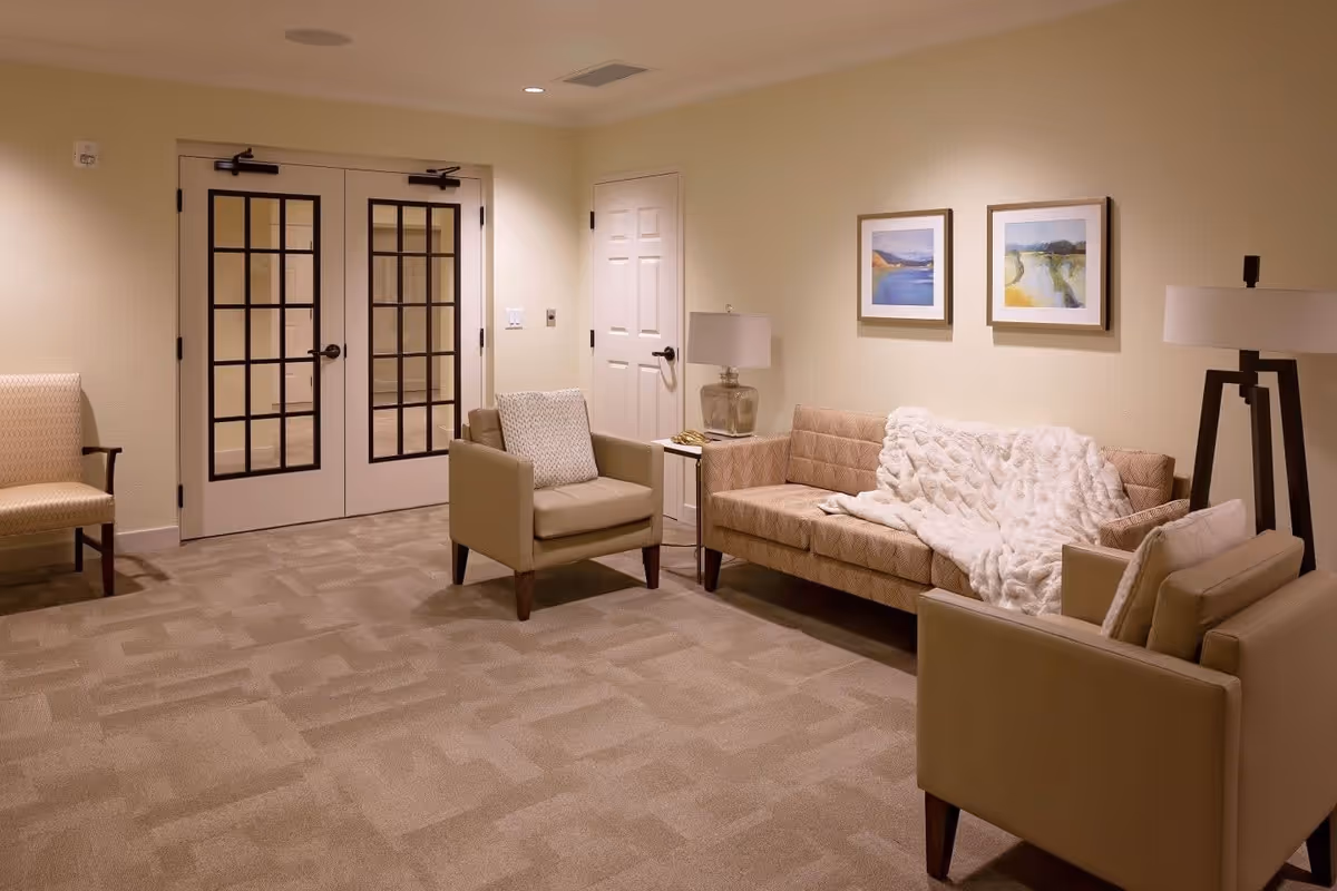 A cozy lounge with a sofa, armchairs, side tables and lamps, framed artwork, and double glass-pane doors.