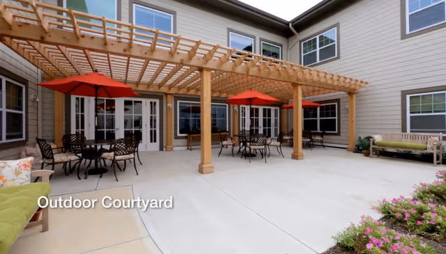 Outdoor courtyard area with a wooden pergola covering several tables and chairs with red umbrellas. The courtyard is surrounded by a beige building with multiple windows and doors. There are benches and some greenery with flowers on the right side.