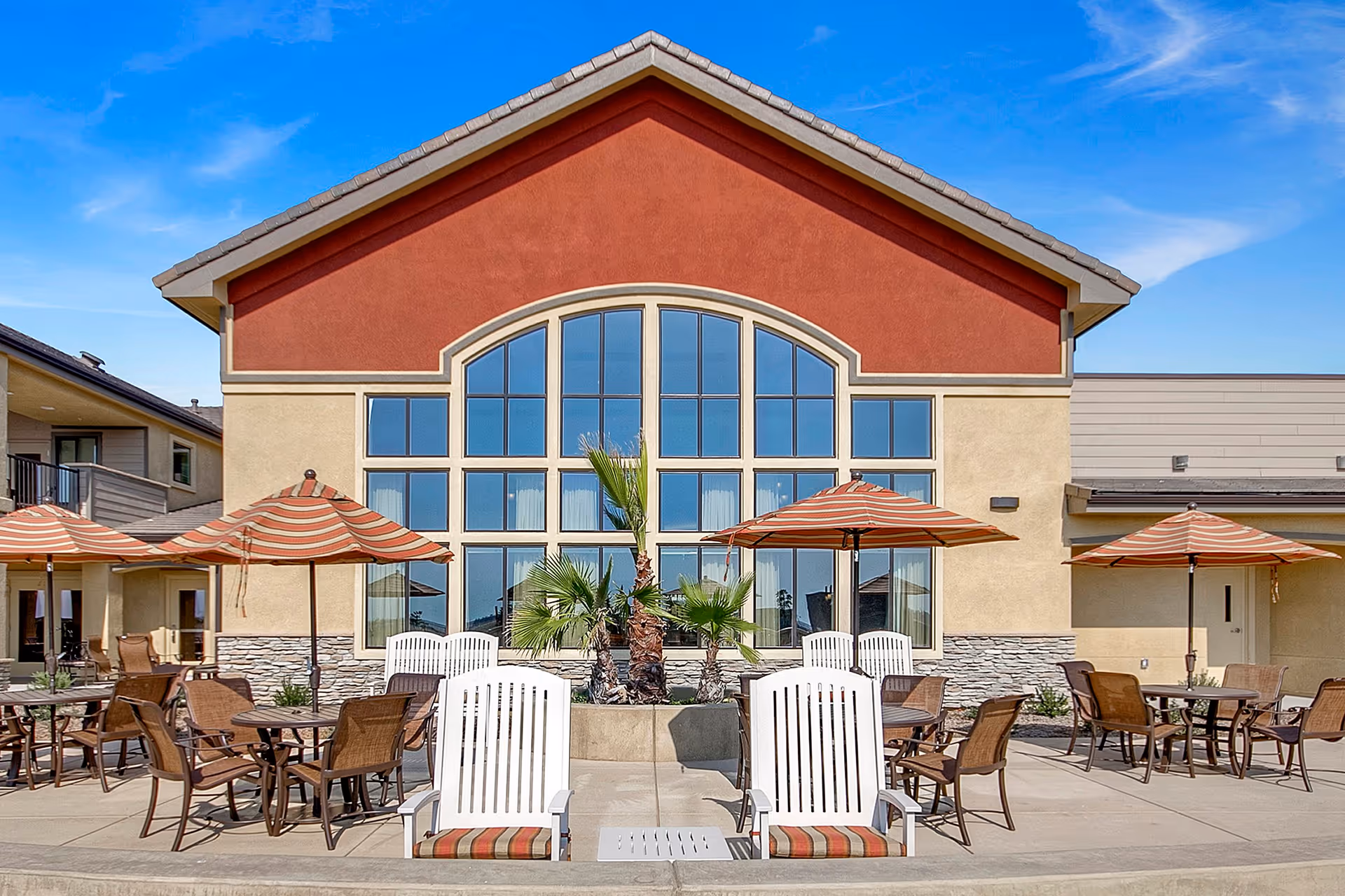 Outdoor patio area of a senior living facility with several tables and chairs under striped umbrellas, white Adirondack chairs in the foreground, and a building with large arched windows and a red and beige exterior in the background under a clear blue sky.