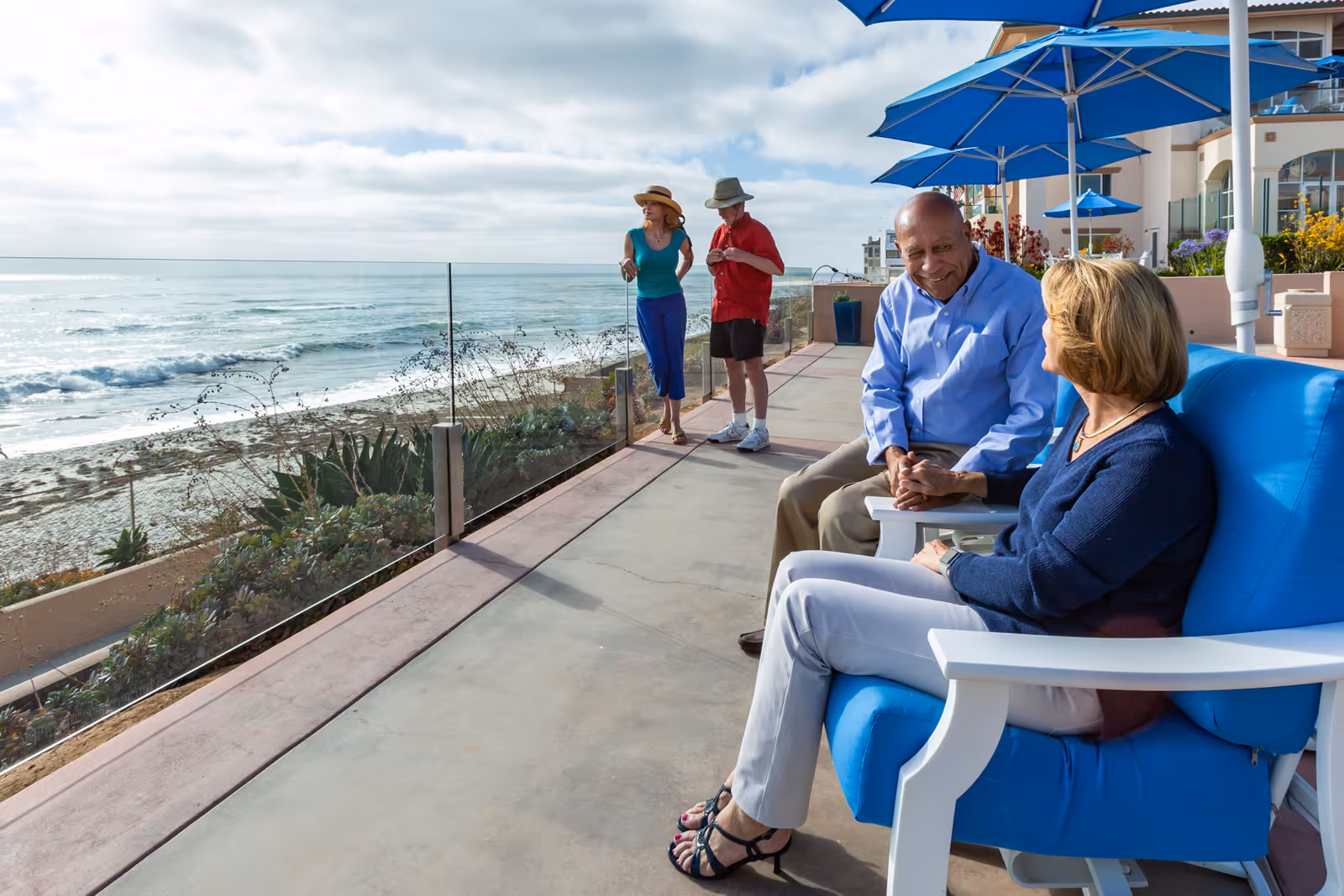 Four older adults relax and chat on a beachfront patio with blue umbrellas and a glass railing overlooking the ocean.