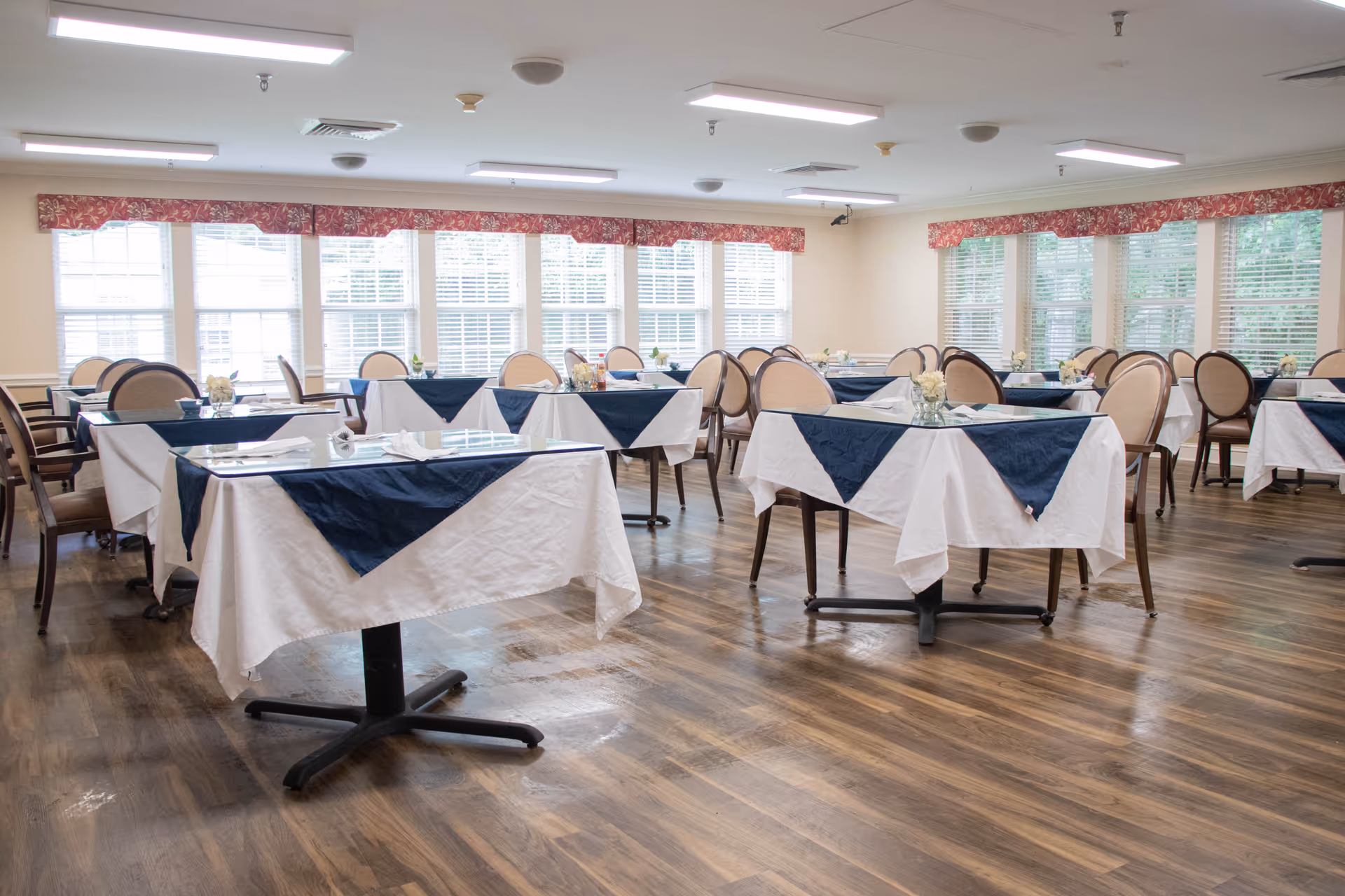 A bright dining room with multiple tables covered in white tablecloths and navy blue overlays. Each table is set with small floral centerpieces and napkins. The room has large windows with white blinds and red patterned valances, and wooden flooring.