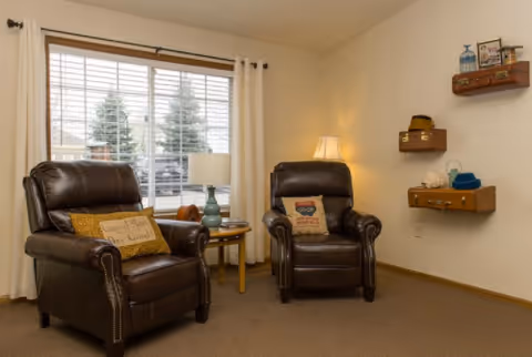 Two brown leather armchairs with throw pillows beside a side table and lamp in a cozy living room with a window and decorative wall shelves.