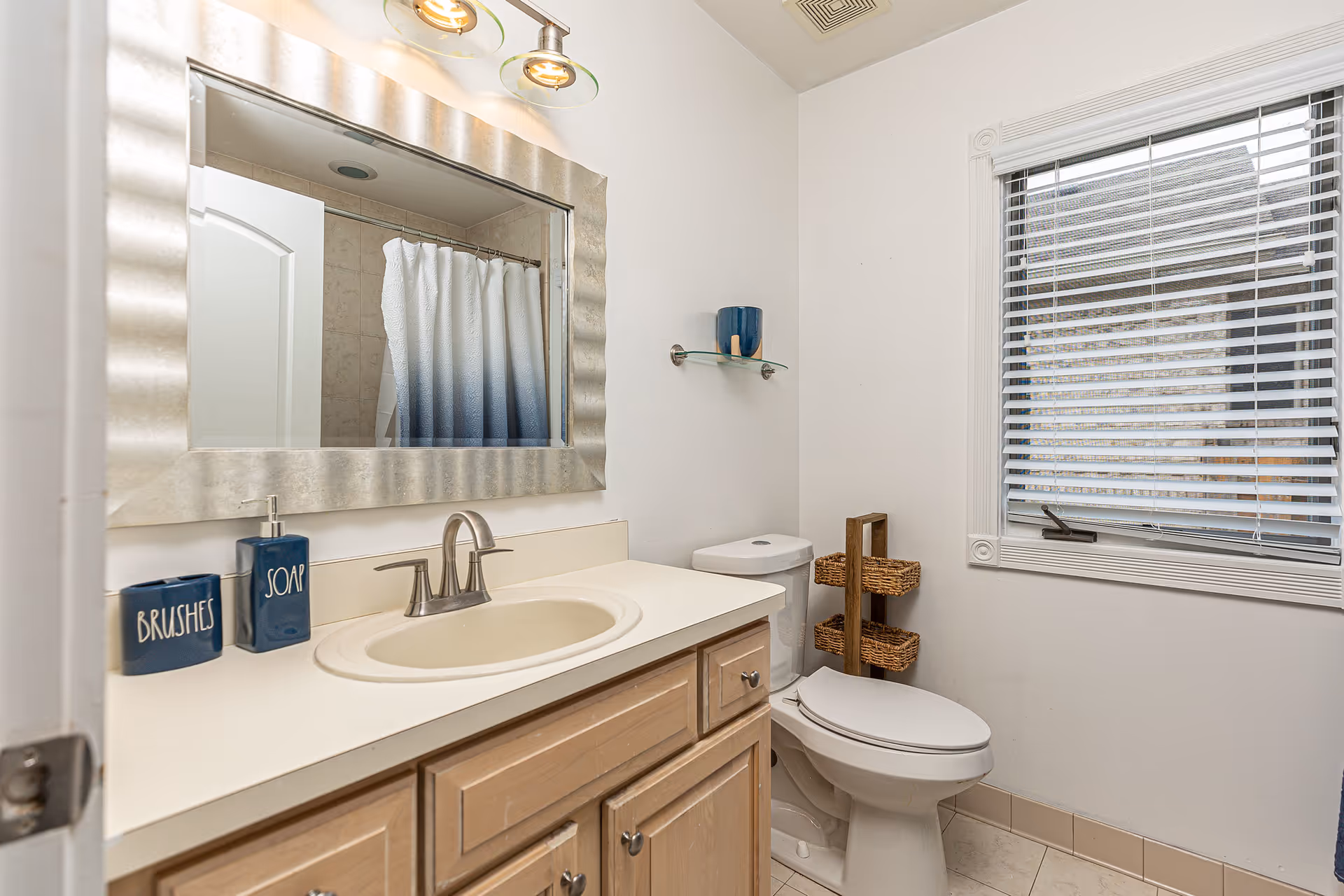 Small bright bathroom featuring a sink vanity with a decorative mirror, a toilet, a window with blinds, and a shower curtain reflected in the mirror.