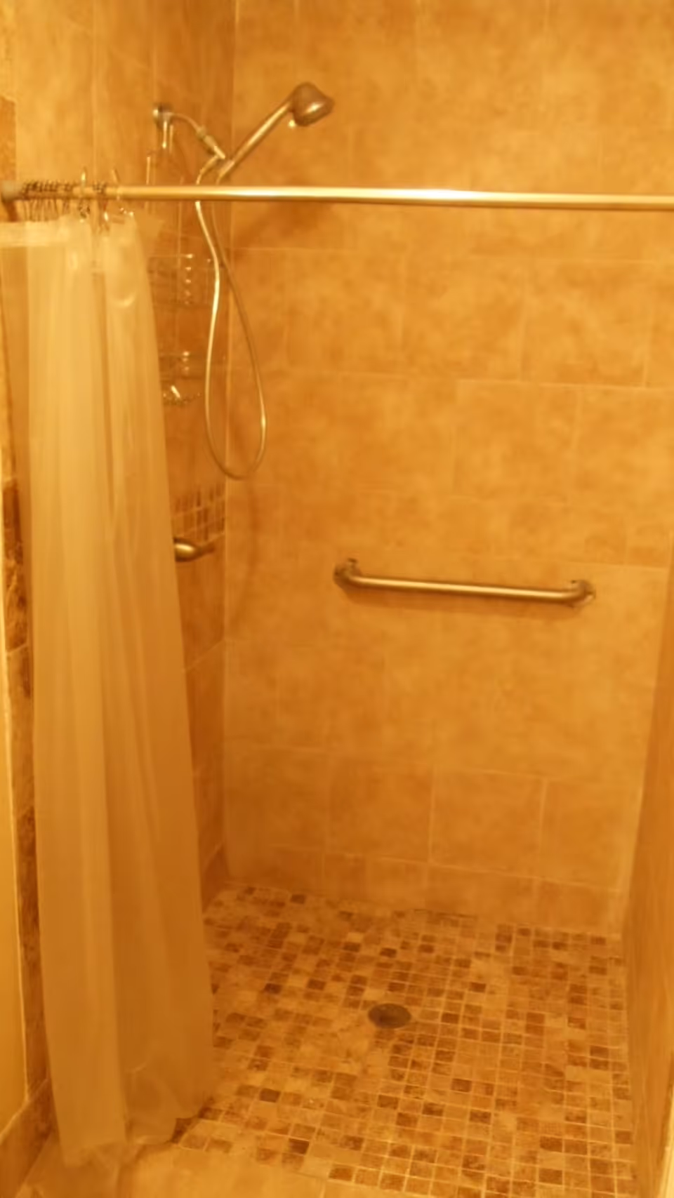 A tiled walk-in shower with a handheld showerhead, a metal grab bar on the wall, and a translucent shower curtain hanging from a curved shower rod.