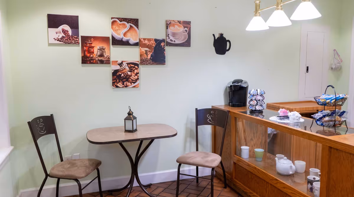 Cozy coffee nook with two chairs and a small table, coffee-themed wall art, and a counter holding a Keurig, cups, and snacks.