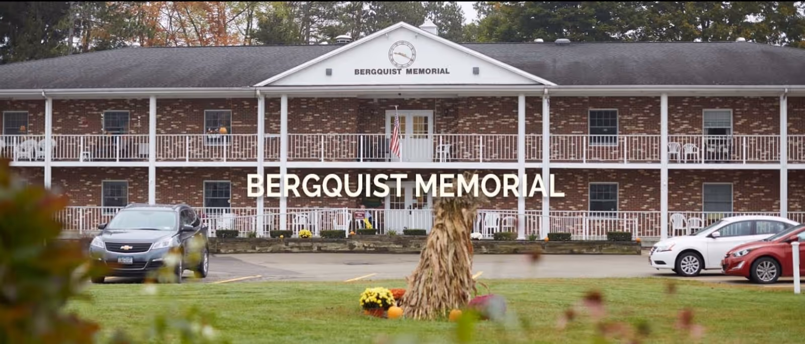 Front exterior view of Bergquist Memorial, a two-story brick building with white railings and columns. There are several cars parked in front and some autumn decorations on the lawn.