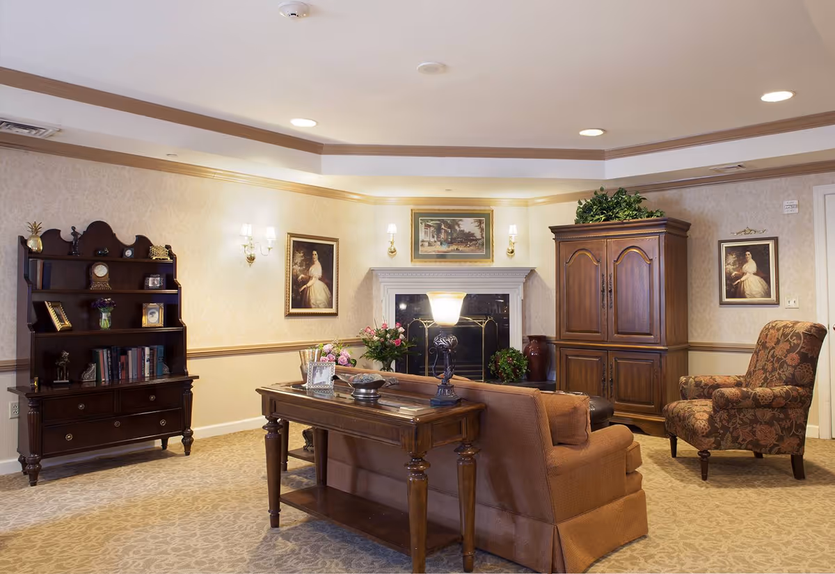A cozy living room area with a brown sofa, a wooden table with a lamp, a floral armchair, a wooden cabinet, and a bookshelf. The room features a fireplace with a painting above it, wall sconces with lights, and framed portraits on the walls. The carpet has a subtle pattern and there are decorative plants on the cabinet and near the fireplace.