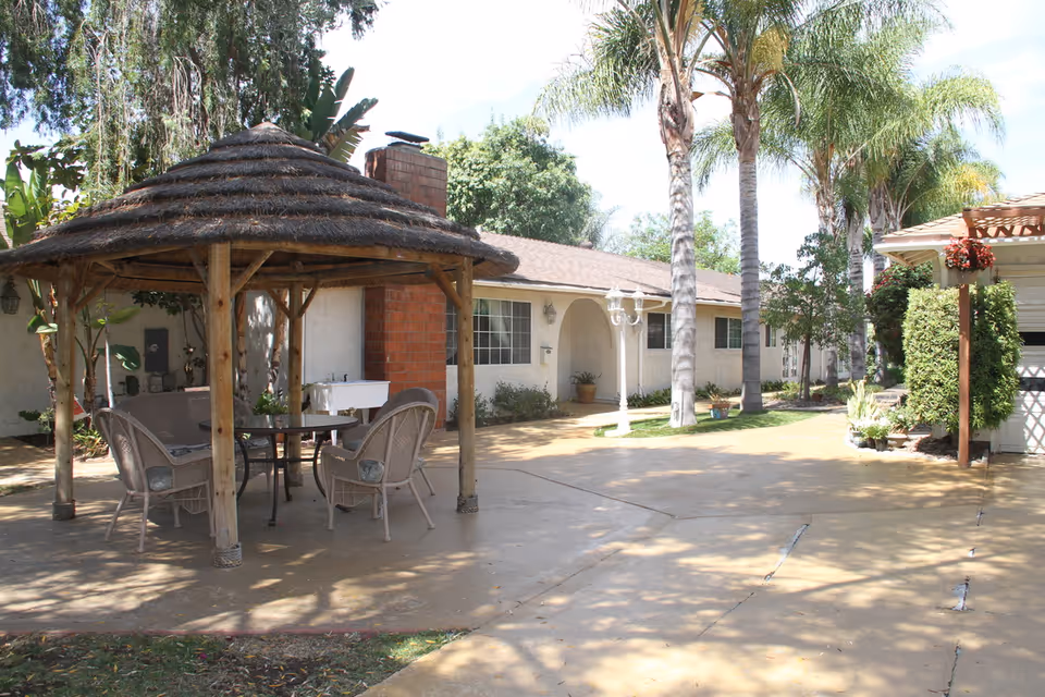 Outdoor patio area with a thatched gazebo covering a round glass table and four wicker chairs. The patio is surrounded by palm trees and other greenery, with a single-story building in the background featuring arched doorways and windows.