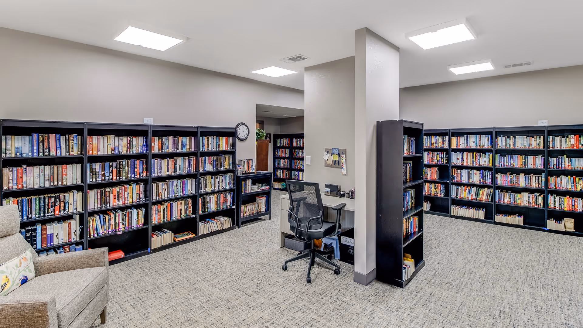 A well-lit library room with multiple black bookshelves filled with books along the walls. There is a gray armchair with a decorative pillow on the left side and a small desk with a black office chair in the center near a column. The room has beige carpet and light gray walls with ceiling lights.