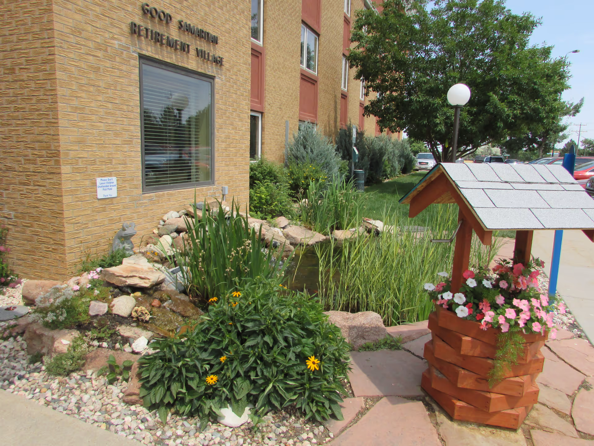 Outdoor garden area at Good Samaritan Retirement Village featuring a small pond surrounded by rocks and plants, a wooden wishing well planter filled with colorful flowers, and a tree near the sidewalk with parked cars in the background.