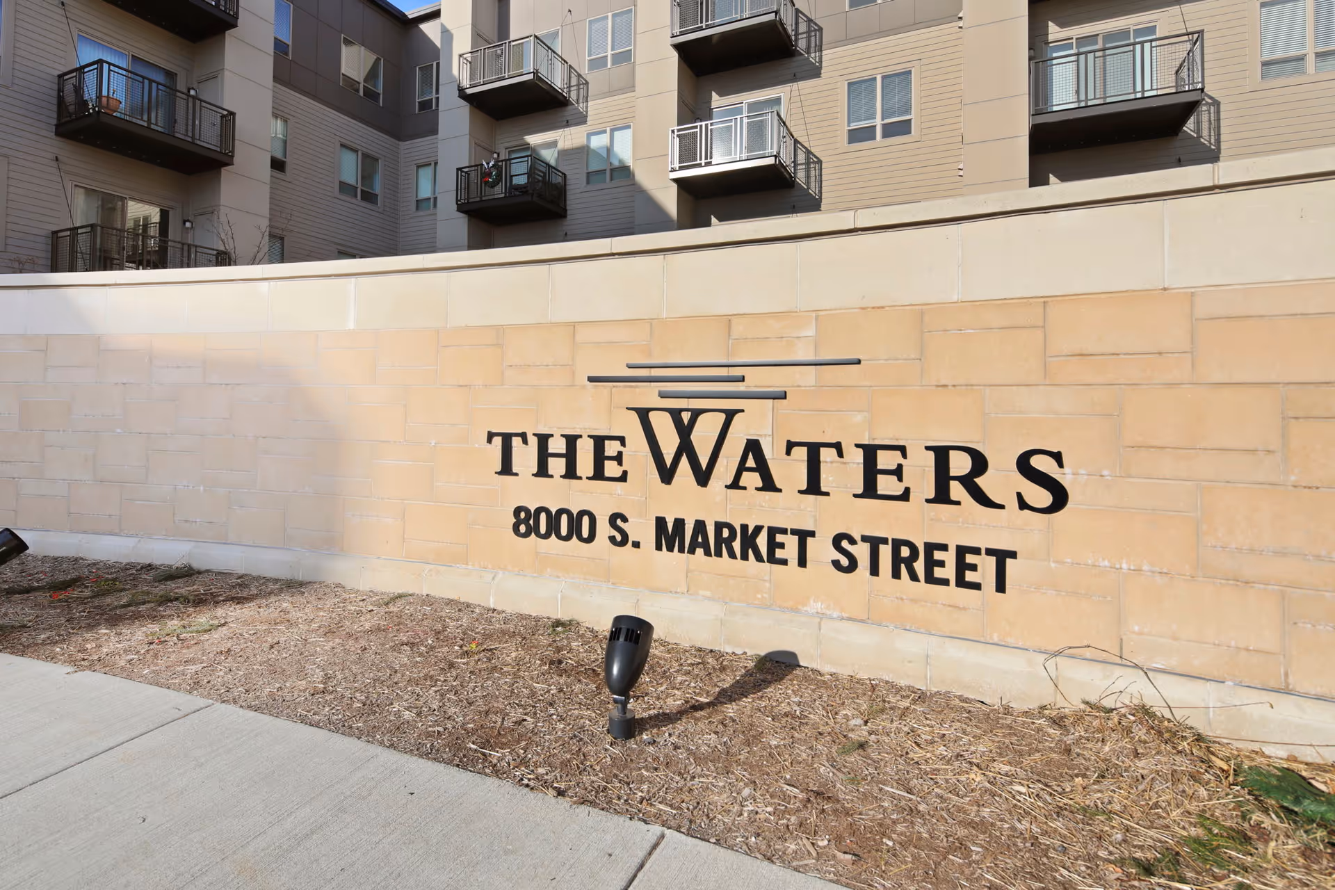 Exterior view of a beige stone wall with the sign 'THE WATERS 8000 S. MARKET STREET' in black letters, with a multi-story residential building with balconies in the background.