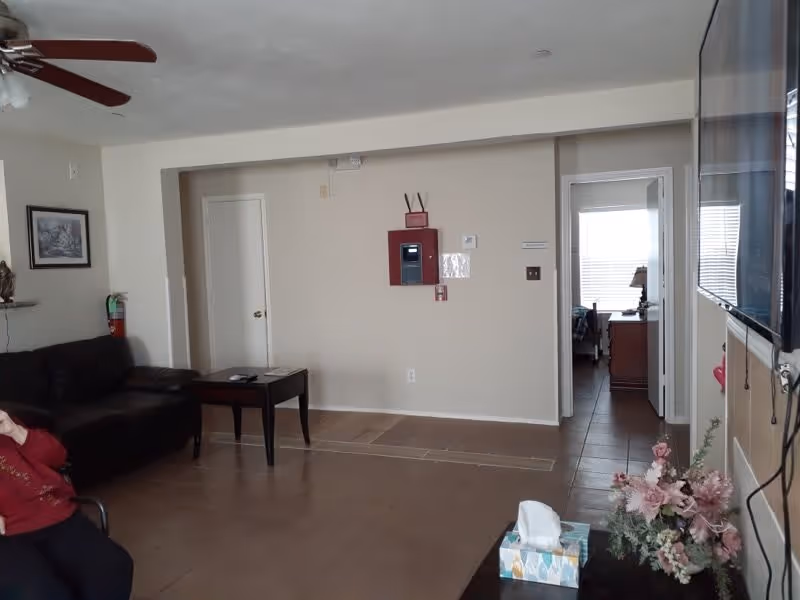 A living room area with a dark brown couch on the left, a small black coffee table in front of it, and a ceiling fan above. There is a hallway with two doorways on the right side, one leading to a room with a bed and a lamp. On the right side of the image, a large flat-screen TV is mounted on the wall above a table with a tissue box and a floral arrangement. The walls are light beige, and the floor is tiled. A fire alarm panel is mounted on the wall in the center background.
