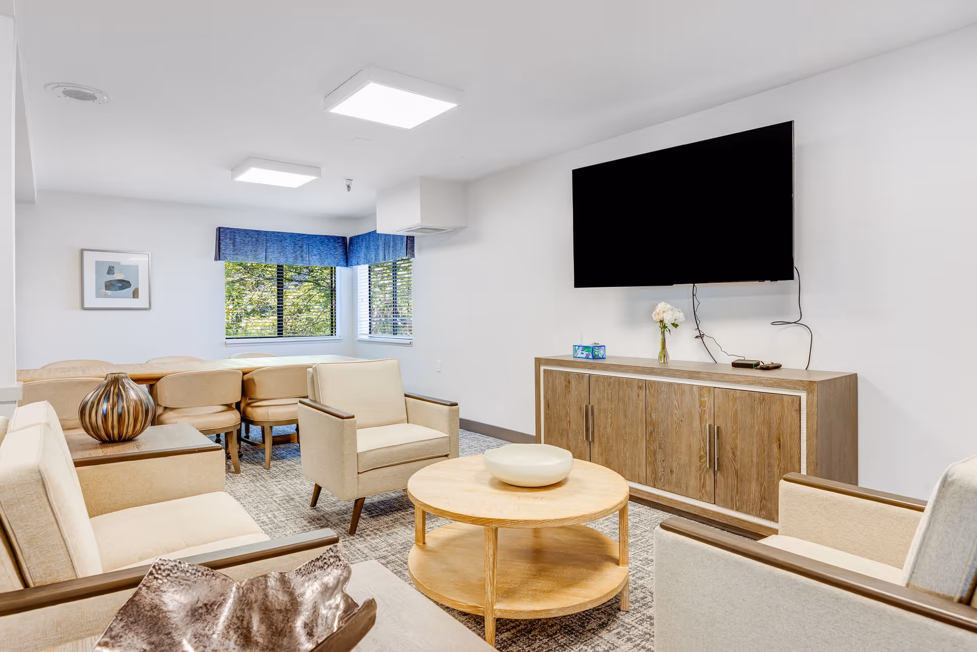 A bright and modern living room area with beige armchairs and a sofa arranged around a round wooden coffee table. A wooden cabinet with a flat-screen TV mounted above it is against the wall. In the background, there is a dining table with several chairs near windows with blue valances, letting in natural light.
