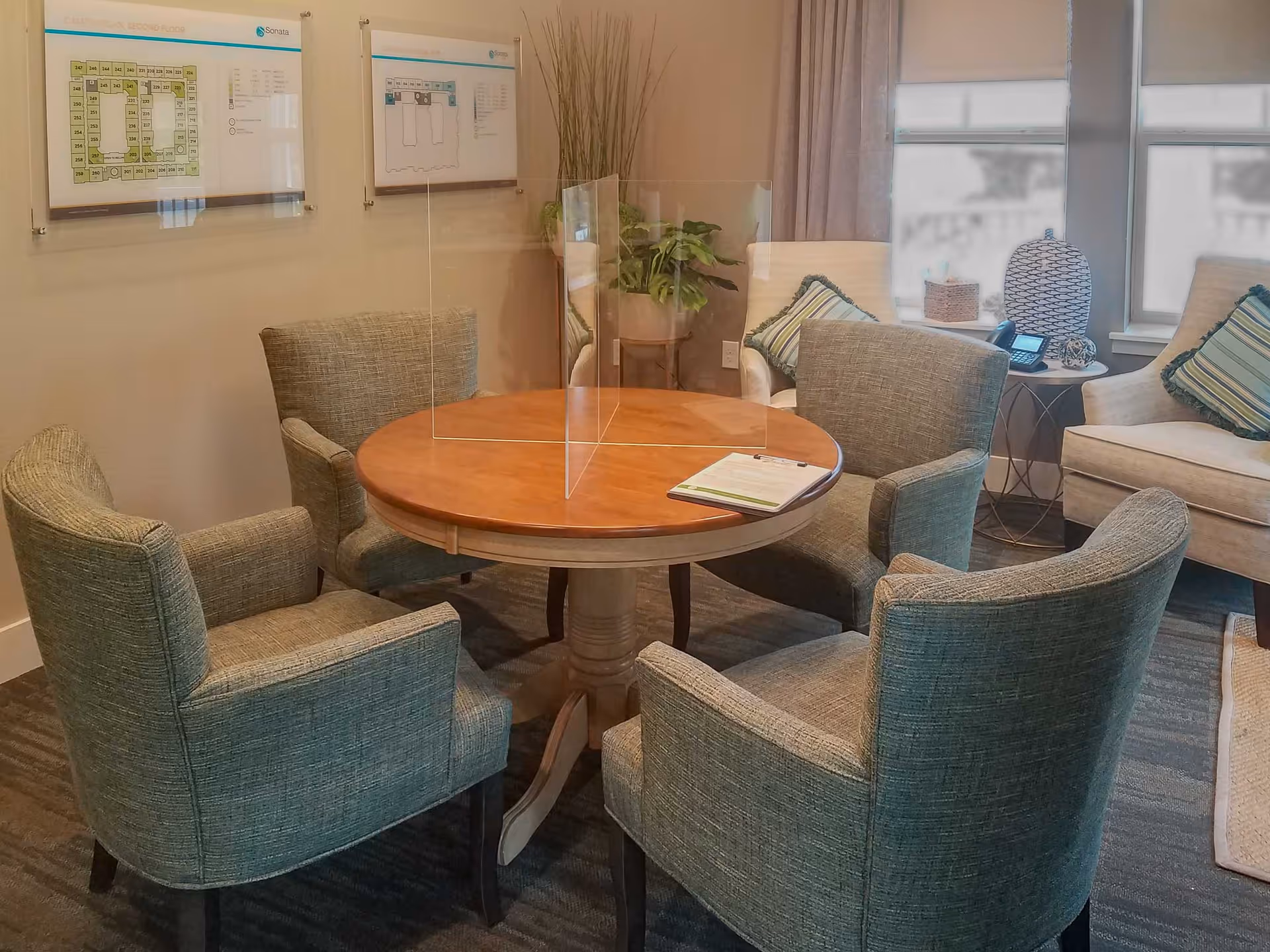 A cozy seating area with a round wooden table surrounded by four upholstered armchairs. A clear acrylic divider is placed on the table. In the background, there are two armchairs with striped cushions, a small side table with a phone and decorative items, and two framed floor plans on the wall.
