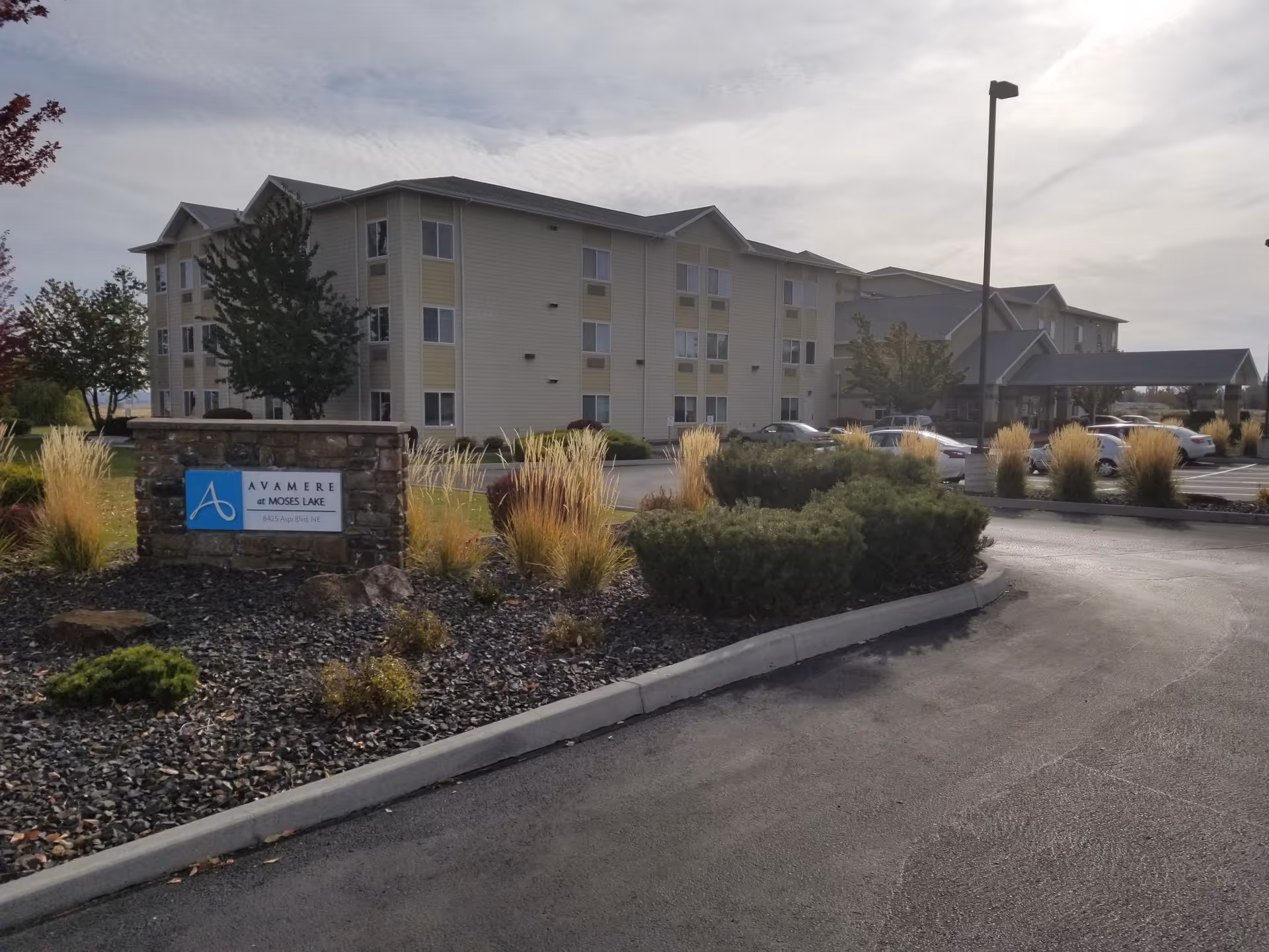 Exterior view of Avamere at Moses Lake, a multi-story senior living facility with a parking lot, landscaping including bushes and ornamental grasses, and a sign displaying the facility's name.