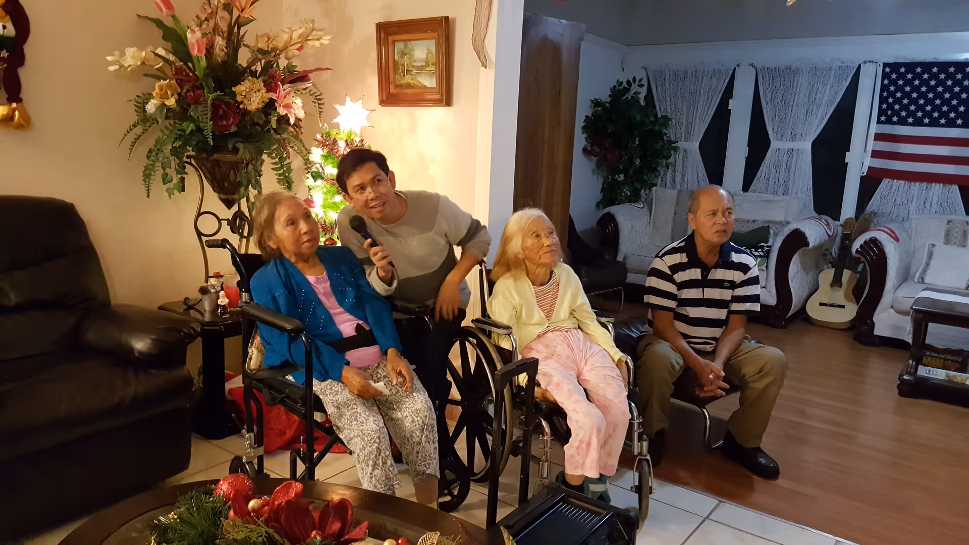 A cozy living room with four people, including two elderly women in wheelchairs, a man holding a microphone, and another man sitting on a chair. The room is decorated with a large floral arrangement, a small Christmas tree, an American flag on the wall, and comfortable seating.