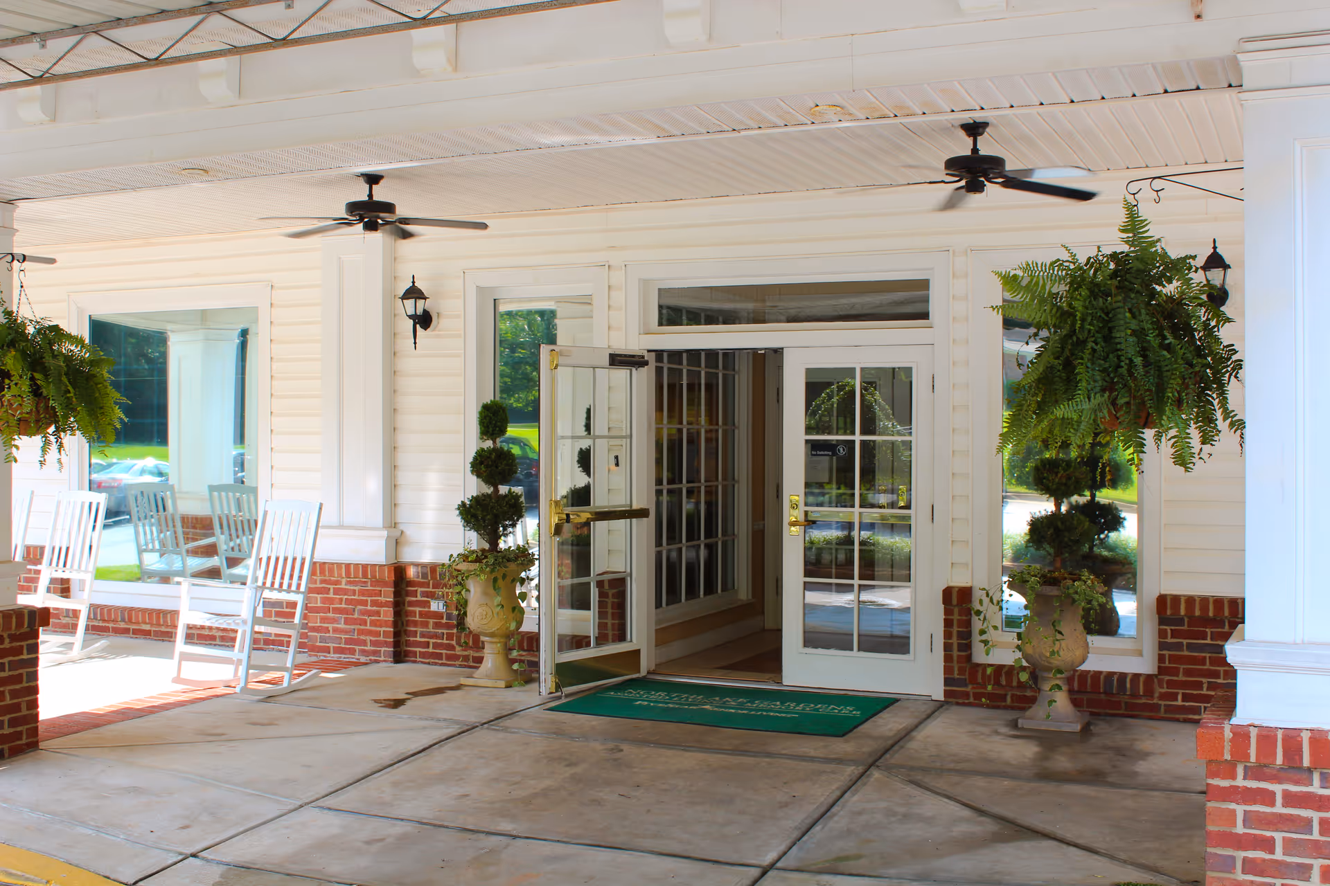 Entrance of a senior living facility with white framed glass doors, two potted plants on either side, hanging ferns, ceiling fans, and white rocking chairs on a brick and concrete porch.