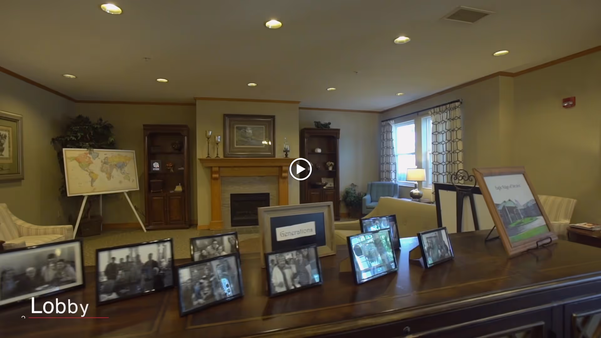 Lobby area of Eagle Ridge of Decatur featuring a wooden table with framed photos, a fireplace with a wooden mantle, two wooden bookshelves, a world map on an easel, and seating area with chairs and a sofa near windows with patterned curtains.