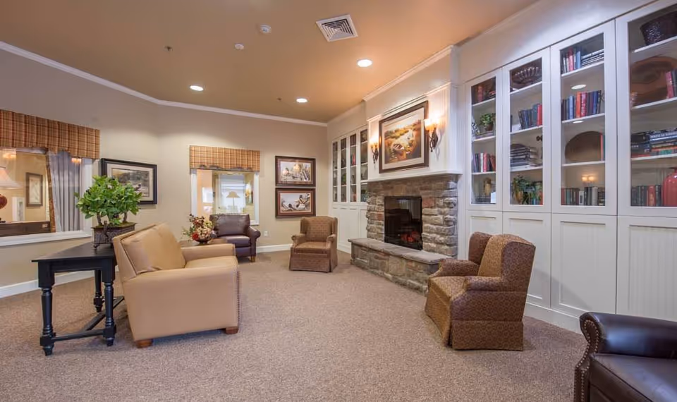 A cozy senior living common area with beige walls and carpeted floor, featuring a stone fireplace with a painting above it, built-in white cabinets with glass doors filled with books and decorative items, and several upholstered armchairs and a leather sofa arranged around the room. There are windows with plaid valances and framed artwork on the walls.