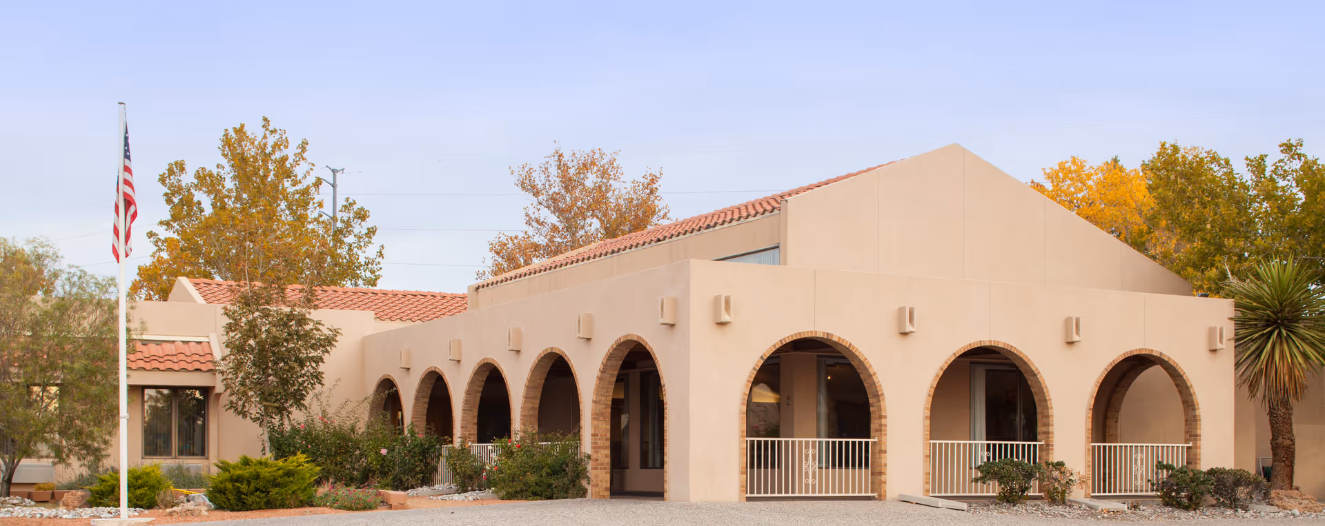 Exterior view of a beige stucco building with a series of arched openings and a tiled roof, surrounded by trees with autumn foliage and desert landscaping. An American flag is displayed on a flagpole in front of the building.