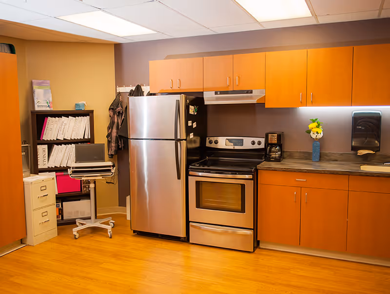 Communal kitchen area with stainless steel refrigerator and oven, wooden cabinets, countertop with coffee maker and vase, and shelving on the left.