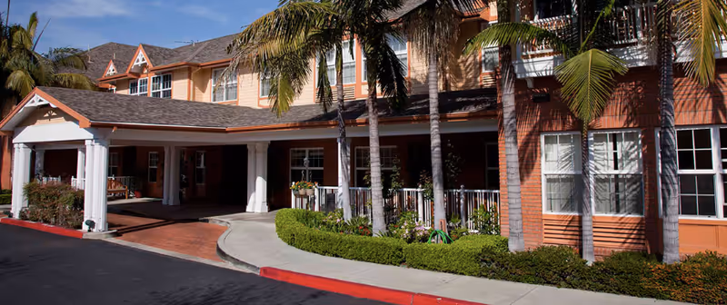 Front entrance of a multi-story senior living building with a covered porte-cochere, palm trees, and landscaped walkway.
