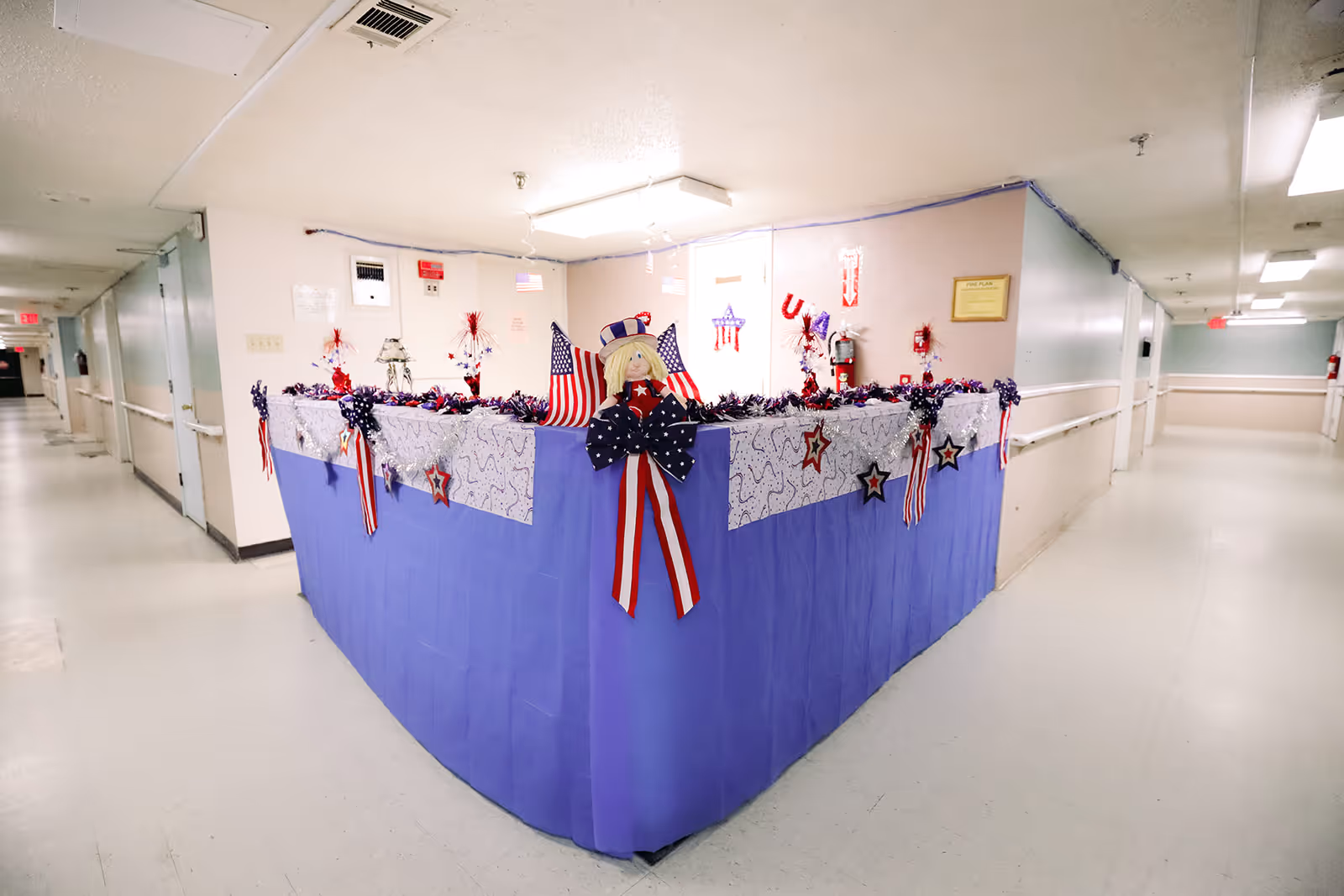 A hallway corner in a healthcare facility decorated with patriotic red, white, and blue decorations including American flags, star ornaments, and a doll wearing a patriotic outfit. The walls and floors are light-colored, and the hallway extends on both sides.