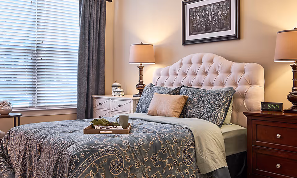 A cozy bedroom with a tufted beige headboard, a bed dressed in blue paisley-patterned bedding, and multiple pillows. There are two bedside tables with lamps on each side of the bed, a digital alarm clock showing 5:44 on the right table, and a window with blinds and dark curtains on the left side. A framed black and white picture hangs on the wall above the bed.