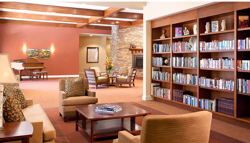 Spacious senior living common area with bookshelves, seating, a coffee table, piano, and a stone fireplace.