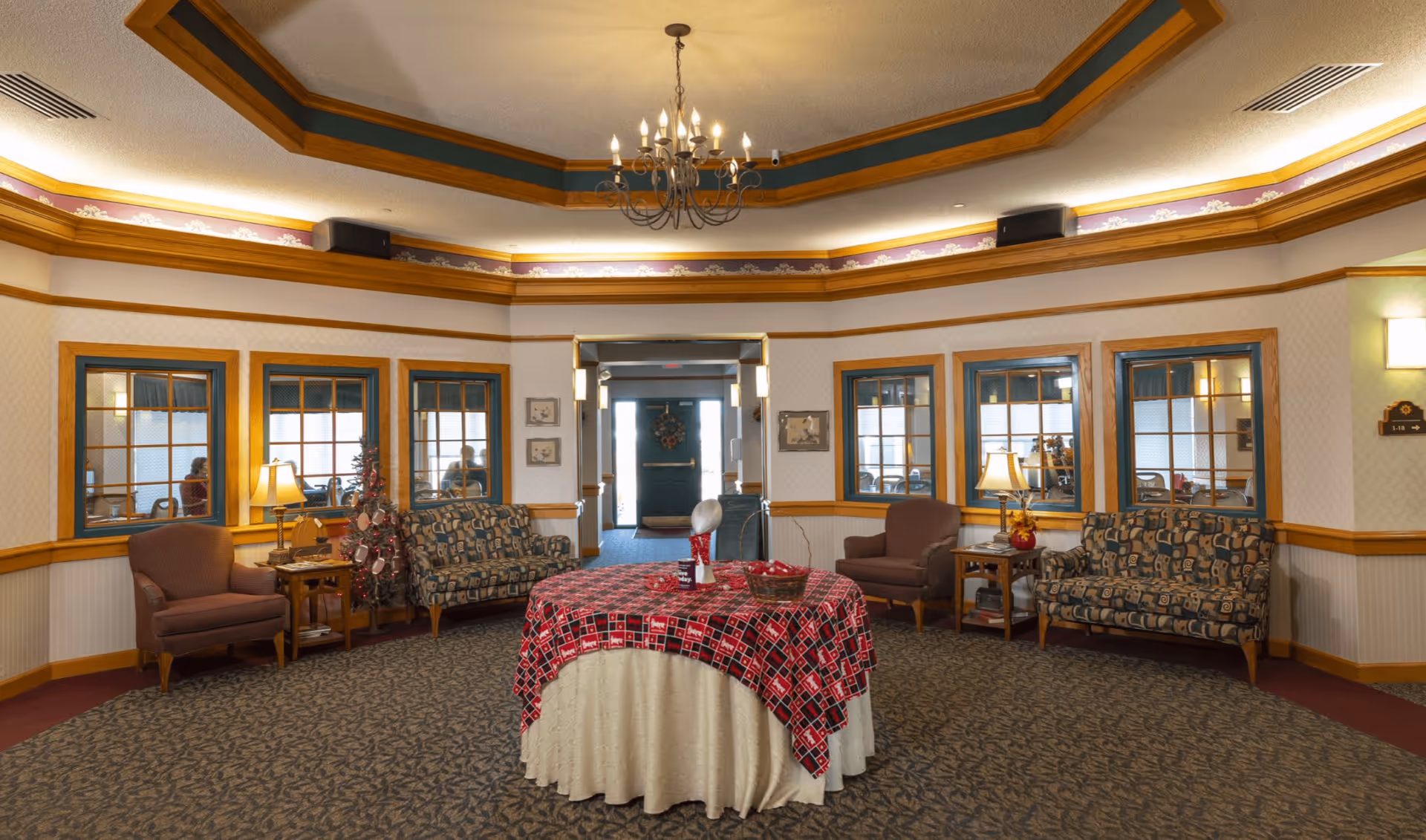 A spacious, well-lit common area with patterned carpet, a round table covered with a red and white tablecloth in the center, surrounded by multiple upholstered chairs and sofas. The walls have wooden trim and several windows looking into adjacent rooms. A chandelier hangs from the ceiling, and lamps on side tables provide additional lighting.