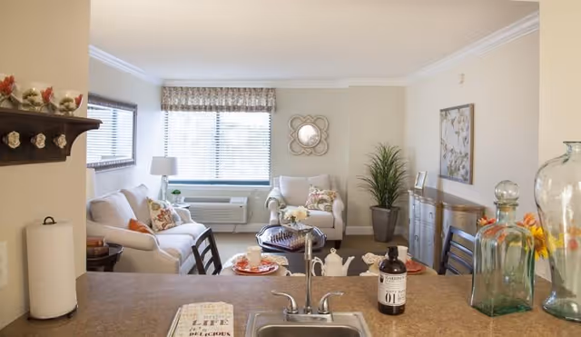 View of a cozy living room from the kitchen counter. The living room features a white sofa and armchair with floral cushions, a coffee table with a chessboard, a large window with blinds and a valance, a decorative mirror on the wall, a tall potted plant, and a sideboard with framed pictures and artwork above it. The kitchen counter in the foreground has a sink, a bottle, a decorative glass container, and a paper towel holder.