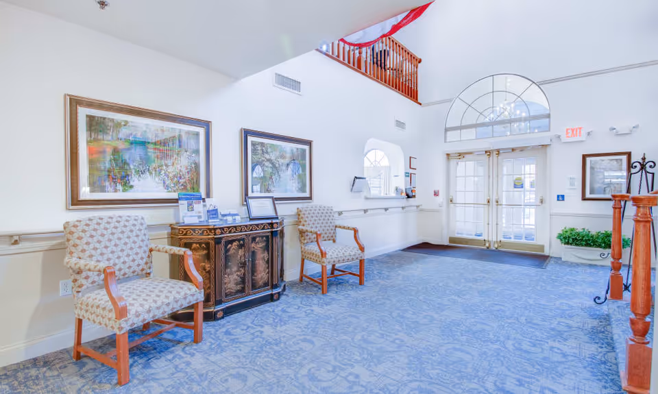 Bright entry lobby with two upholstered chairs, an ornate cabinet, framed paintings, blue patterned carpet and double glass doors.