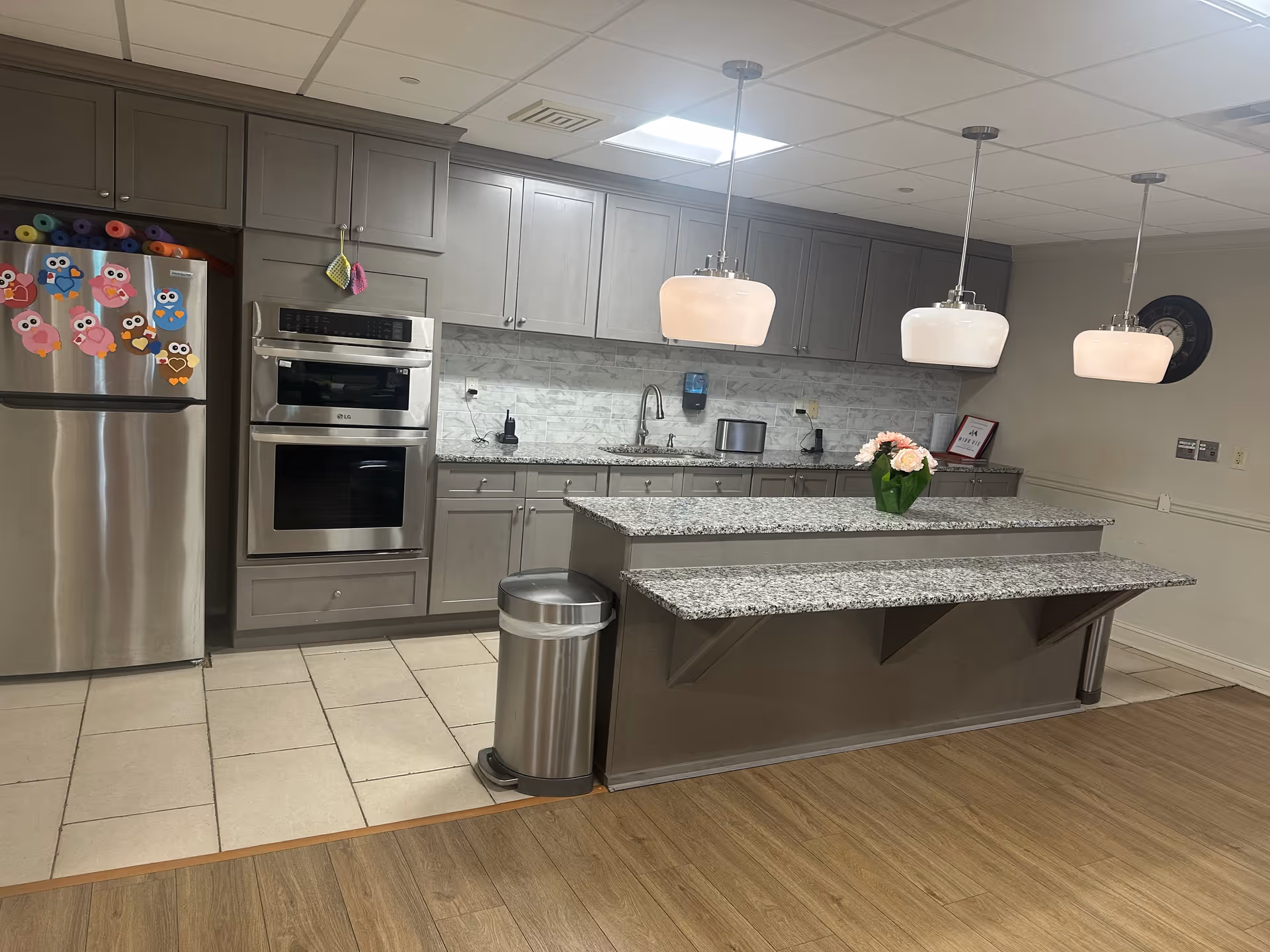 Modern kitchen area with gray cabinets, stainless steel refrigerator decorated with colorful owl magnets, built-in double oven, granite countertops, a kitchen island with a flower vase, three hanging pendant lights, and a wall clock.