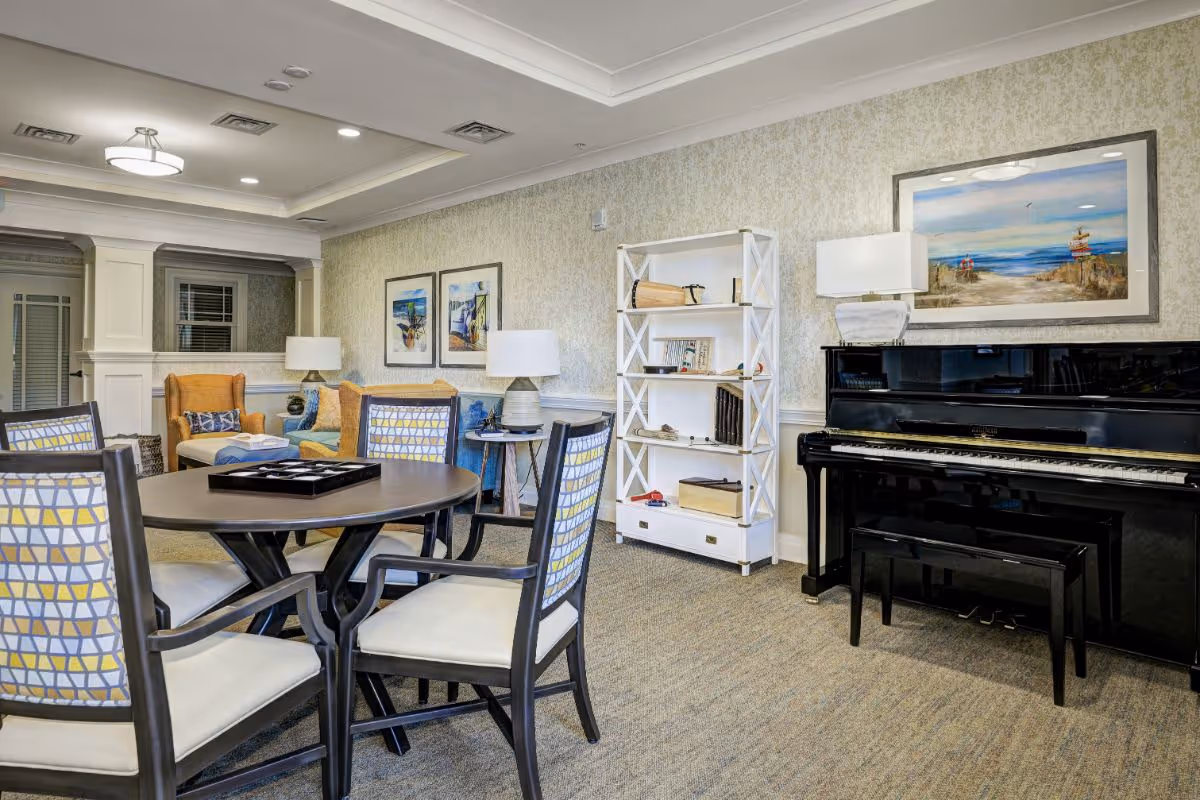 Bright communal lounge with a round table and chairs, upholstered seating, a bookshelf, and a black upright piano.