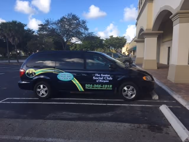 A black minivan parked in a parking lot next to a building with arches. The van has signage on the side that reads 'Adult Day Care Center' and 'The Senior Social Club of Margate' along with a phone number and website.