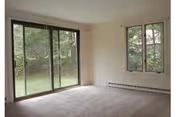 Empty carpeted living room with a large sliding glass door and a window overlooking a green yard.