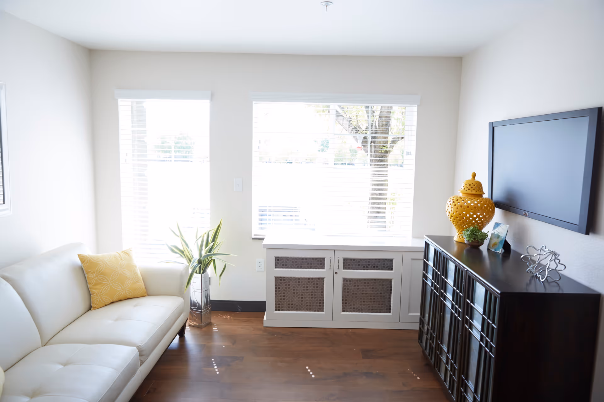 A bright living room with a white leather sofa adorned with a yellow pillow, a potted plant next to the sofa, two large windows with white blinds letting in natural light, a white cabinet beneath the windows, and a dark wooden cabinet with decorative items including a yellow vase, a small plant, a framed photo, and a metal sculpture. A flat-screen TV is mounted on the wall above the dark cabinet.