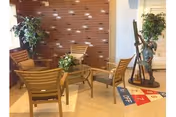 A cozy seating area with four wooden chairs arranged around a glass-top round table, set against a brick wall with small decorative lights. There are two potted plants on either side of the seating area and a shuffleboard game on the floor nearby.