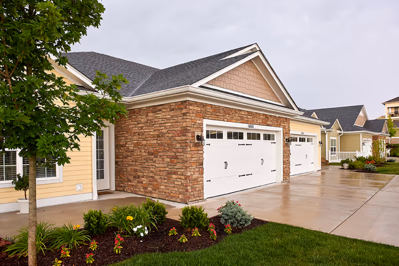 Exterior view of a senior living facility showing a row of attached garages with white doors and stone and yellow siding. There is a concrete driveway, a small landscaped garden with flowers and shrubs, and a tree on the left side under an overcast sky.