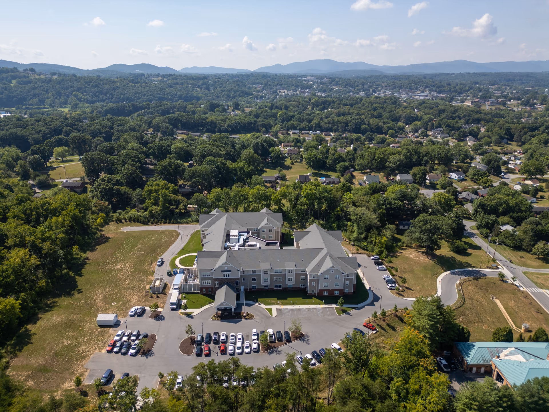 Aerial view of a large senior care facility and parking lot surrounded by trees, nearby houses, and distant mountains.