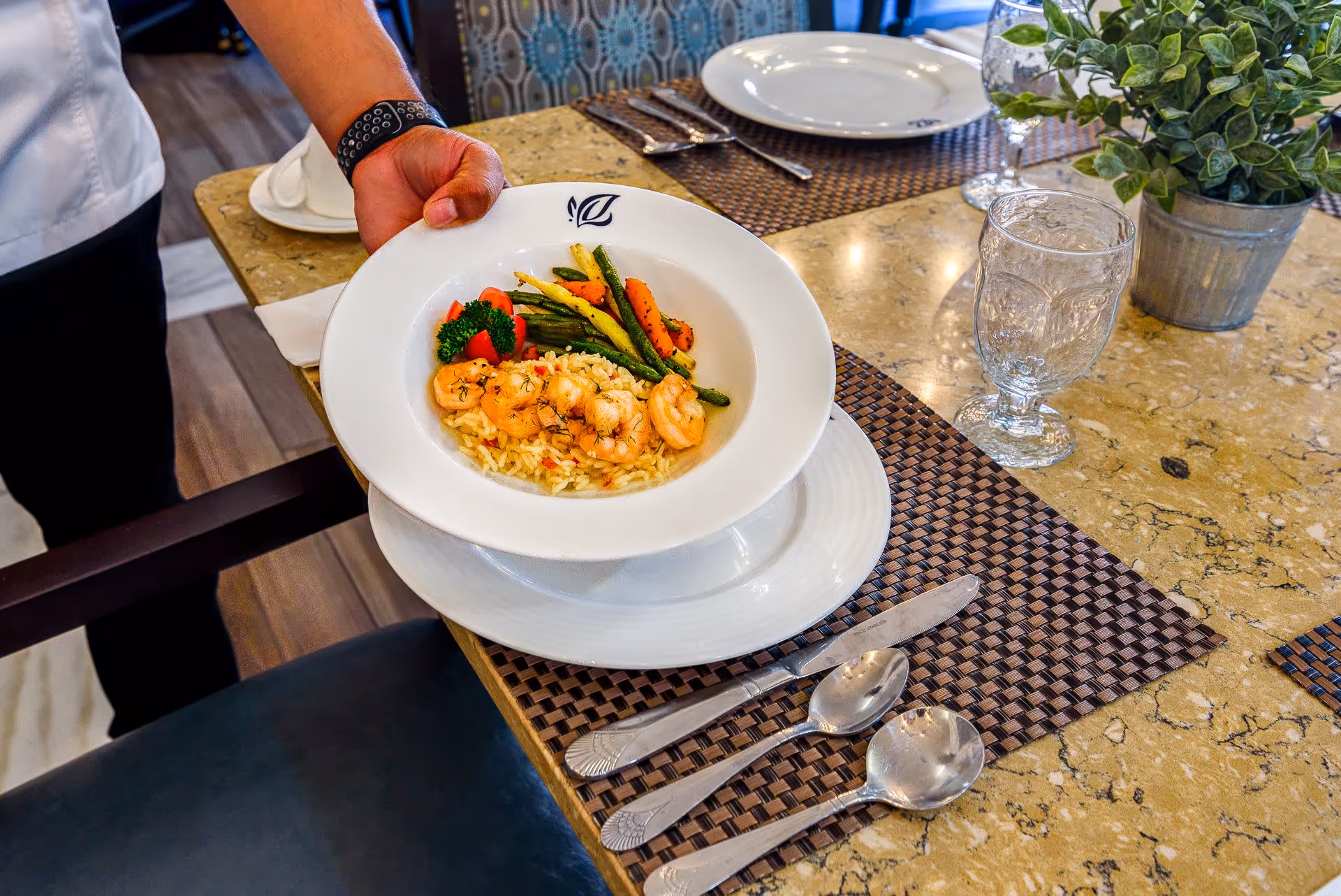 A hand holding a white plate with a meal consisting of shrimp, rice, and mixed vegetables over a table set with silverware, a glass, and a small potted plant.